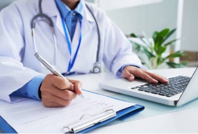 Médico em consultório escrevendo em uma prancheta e usando um laptop.