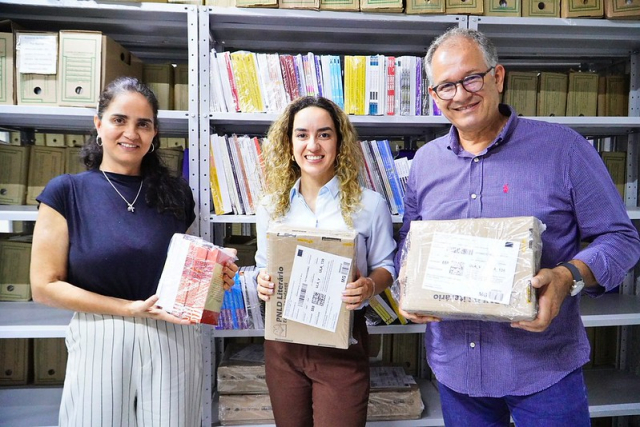Três pessoas posam com pacotes de livros e documentos em uma sala com estantes de arquivos e publicações.