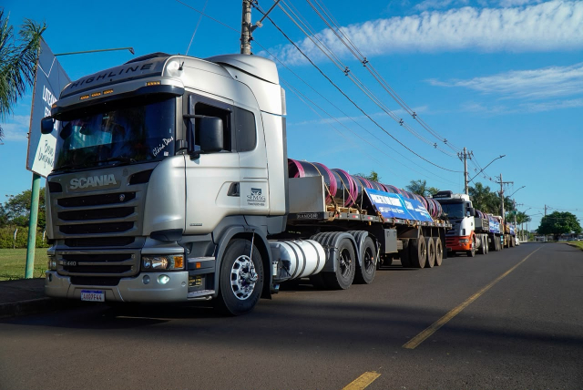Caminhões de carga enfileirados em uma rodovia urbana sob um céu ensolarado.
