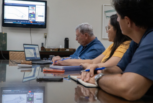 A imagem mostra pessoas sentadas ao redor de uma mesa de conferência, participando de uma reunião. 