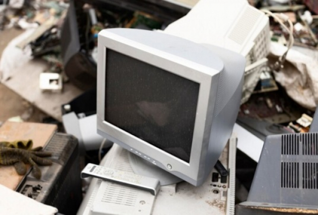 A imagem mostra um monitor CRT antigo descartado em meio a outros equipamentos eletrônicos, possivelmente em um local de descarte de lixo eletrônico. 