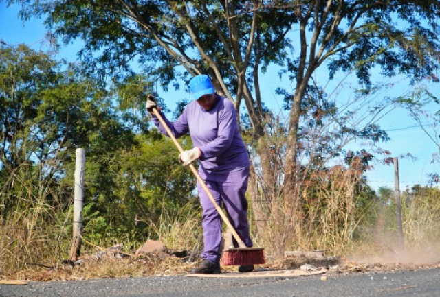 Servidor realiza varrição e limpeza da margem de uma via asfaltada.