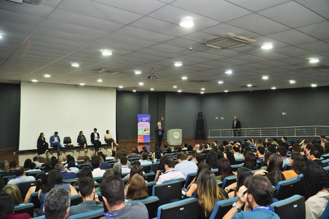 Palestrantes participam de um evento em auditório lotado, com público acompanhando as apresentações.